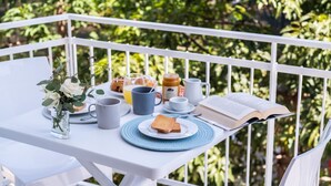 Uma mesa branca com um livro, um prato com torradas, uma xícara, uma caneca e um pote de mel numa varanda com guarda-corpo branco e vegetação ao fundo.