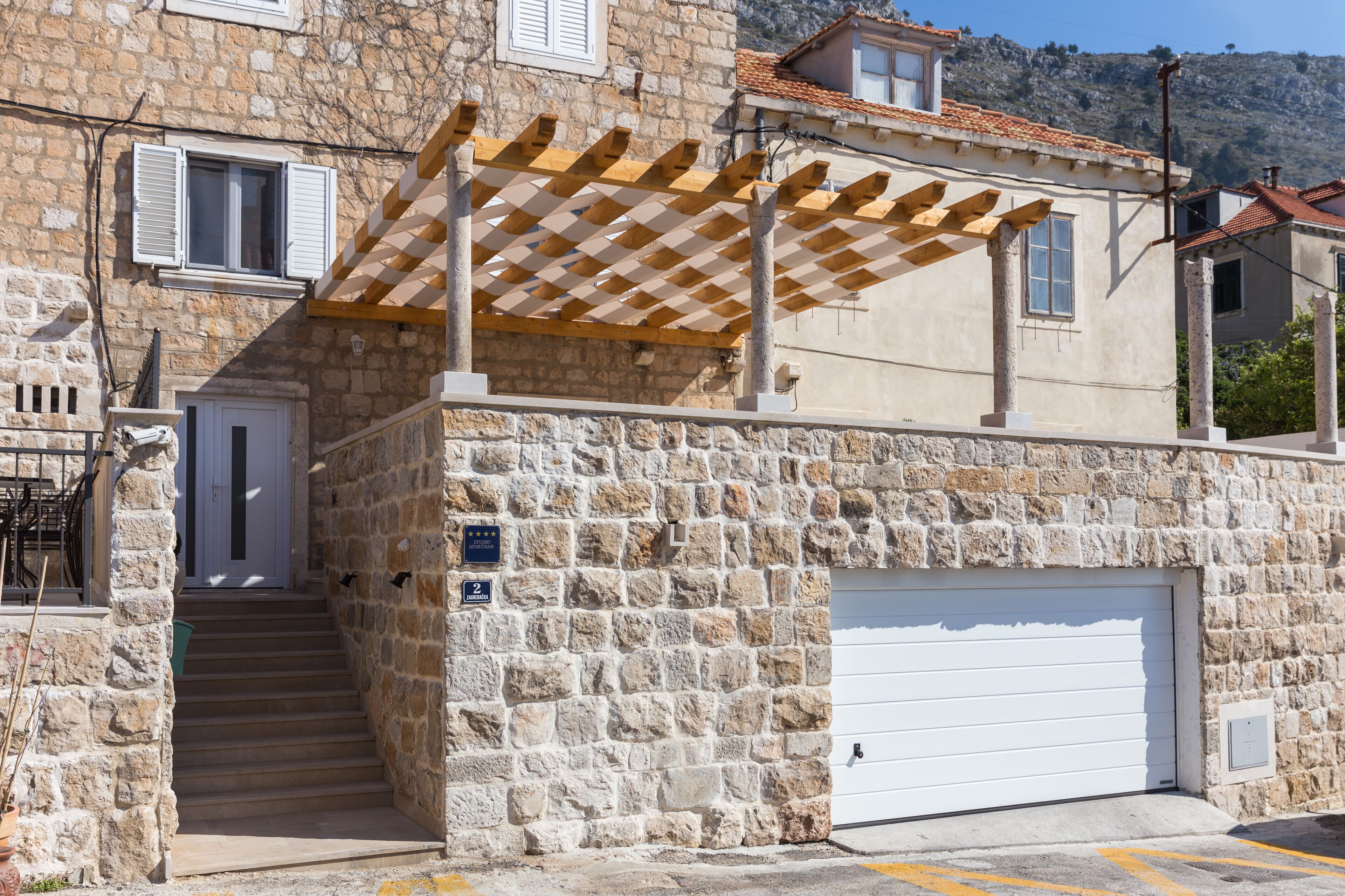 Ein Steinhaus mit weißer Garagentür, einer Holzpergola und einer Treppe, die zum Eingang führt.