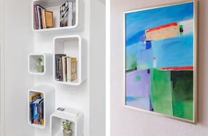 Una estantería moderna con libros y objetos decorativos, una planta pequeña y un cuadro abstracto en la pared.