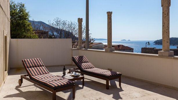 A balcony with two striped lounge chairs, a small table, and a bottle of wine. The balcony overlooks a body of water and ancient ruins. - Apartment Toma (Dubrovnik)