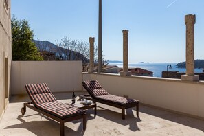 A balcony with two striped lounge chairs, a small table, and a bottle of wine. The balcony overlooks a body of water and ancient ruins.