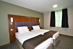 A hotel room with two beds, a wooden headboard, a nightstand, and a window with black curtains. - The Oakwood Hotel (Gloucester)
