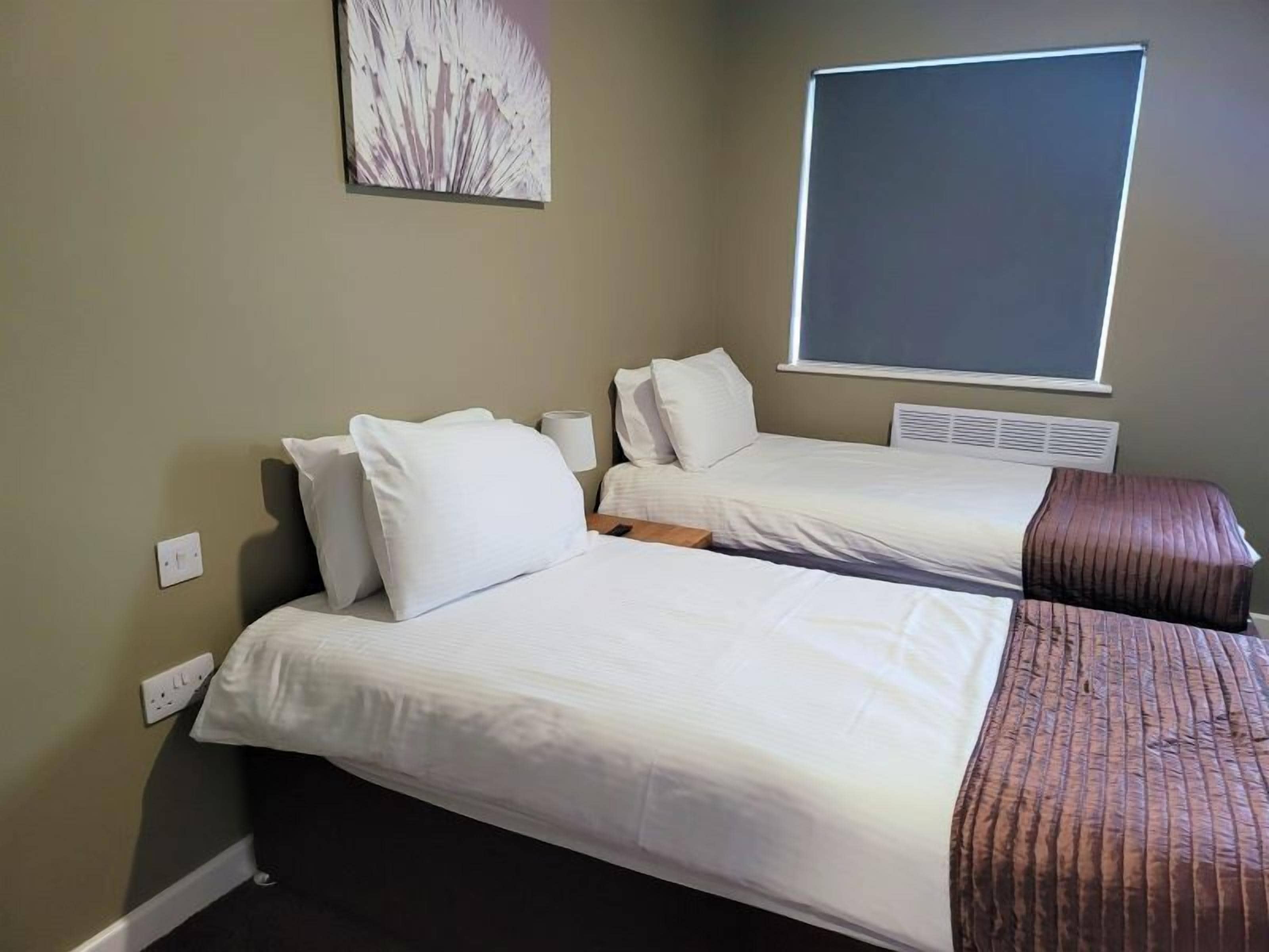 A hotel room with two single beds, a window with blinds, a wall-mounted air conditioning unit, and a power outlet.