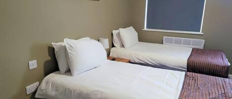A hotel room with two single beds, a window with blinds, a wall-mounted air conditioning unit, and a power outlet.
