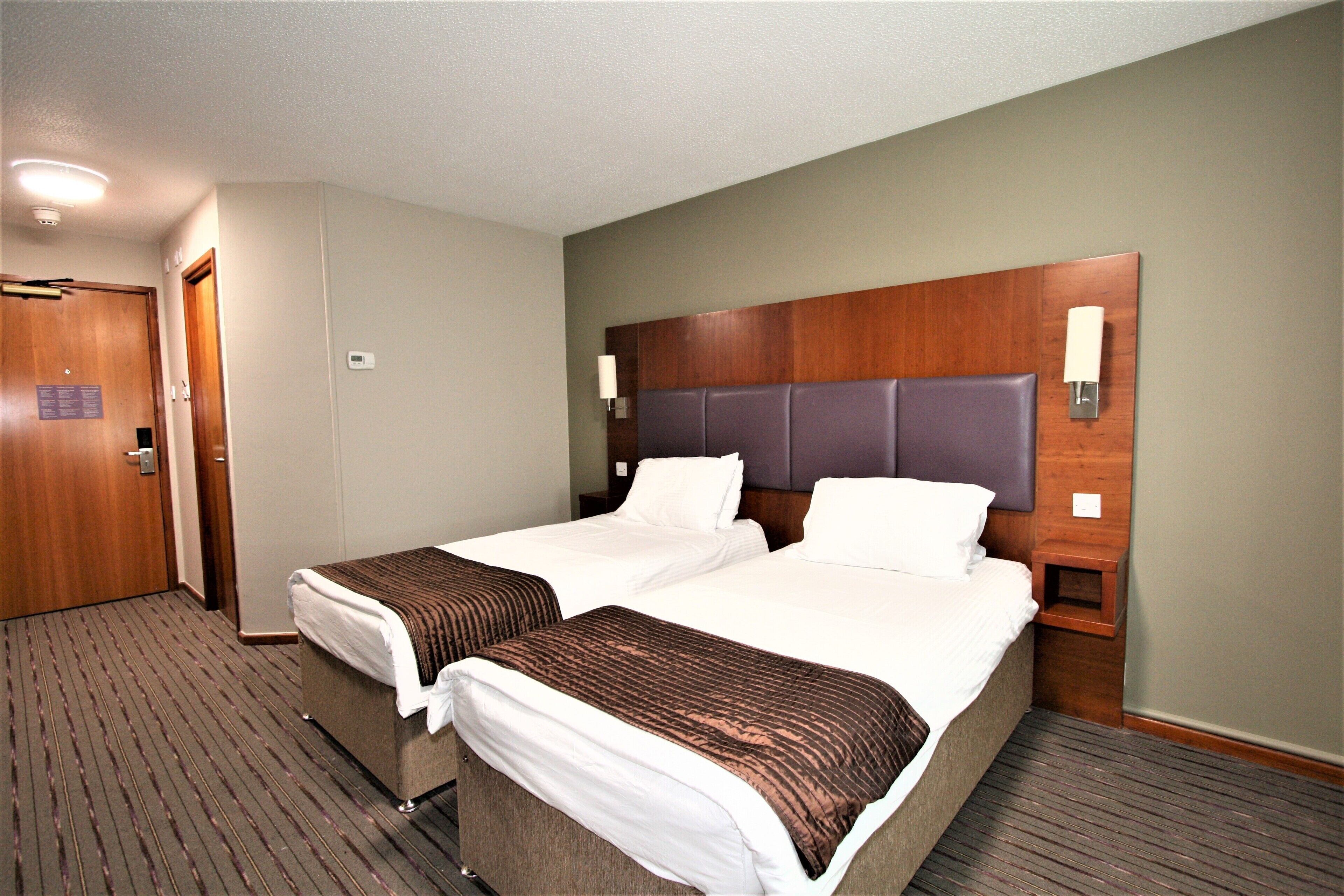A hotel room with two beds, a wooden headboard, and a nightstand with a lamp.