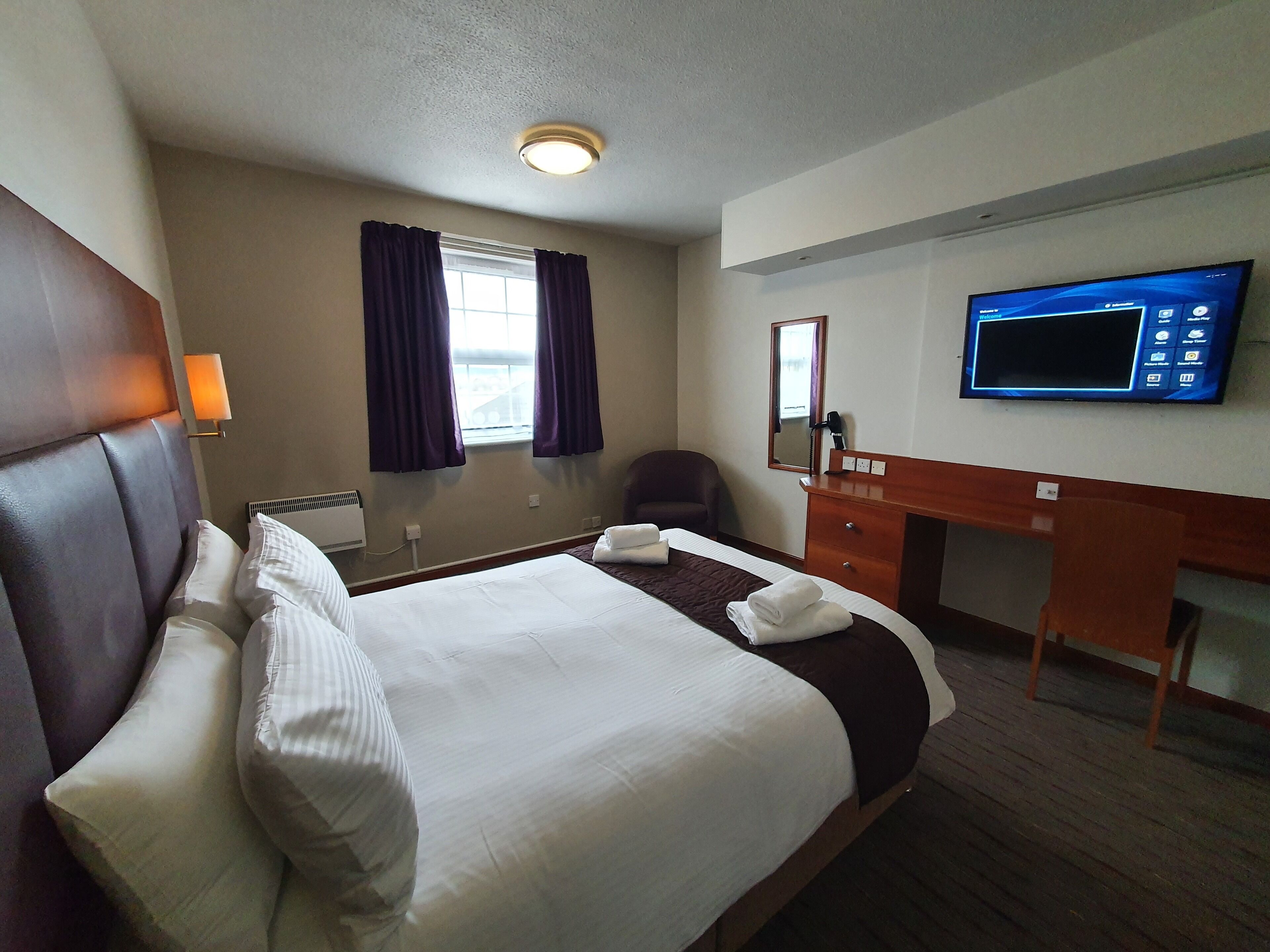 A hotel room with a large bed, a desk with a flat-screen TV, a chair, a desk lamp, and a window with curtains.
