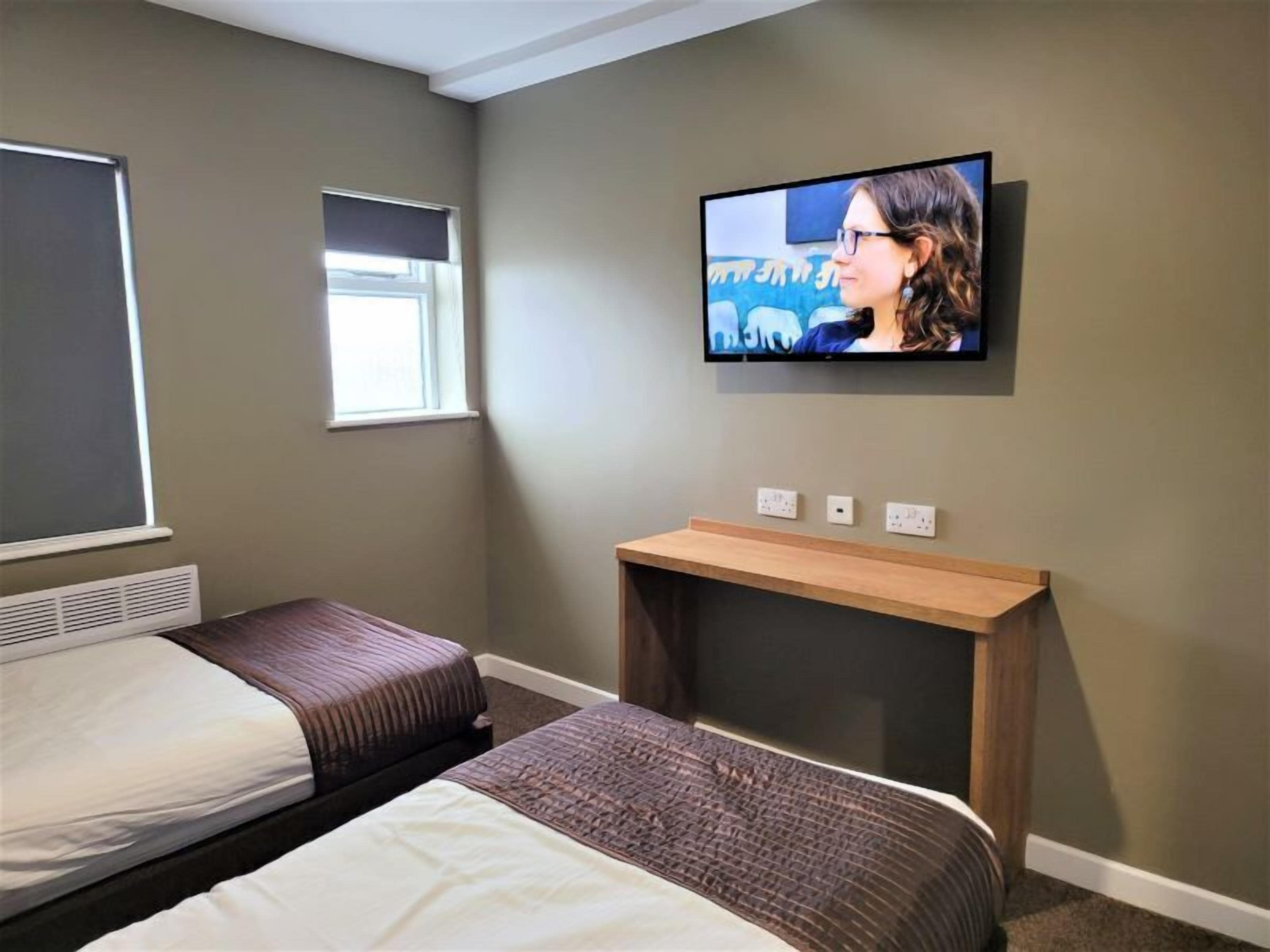 Camera d'albergo con due letti, una TV fissata al muro, una finestra e un piccolo tavolino in legno.