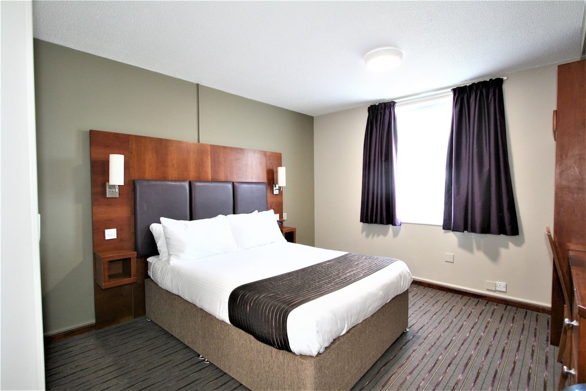 A hotel room with a large bed, wooden headboard, two bedside tables, a nightstand, a window with curtains, and a carpeted floor.