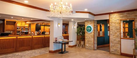 Un vestíbulo de hotel con un mostrador de recepción, una araña de cristal, una zona de bar y un cartel que dice 'The Grill'.