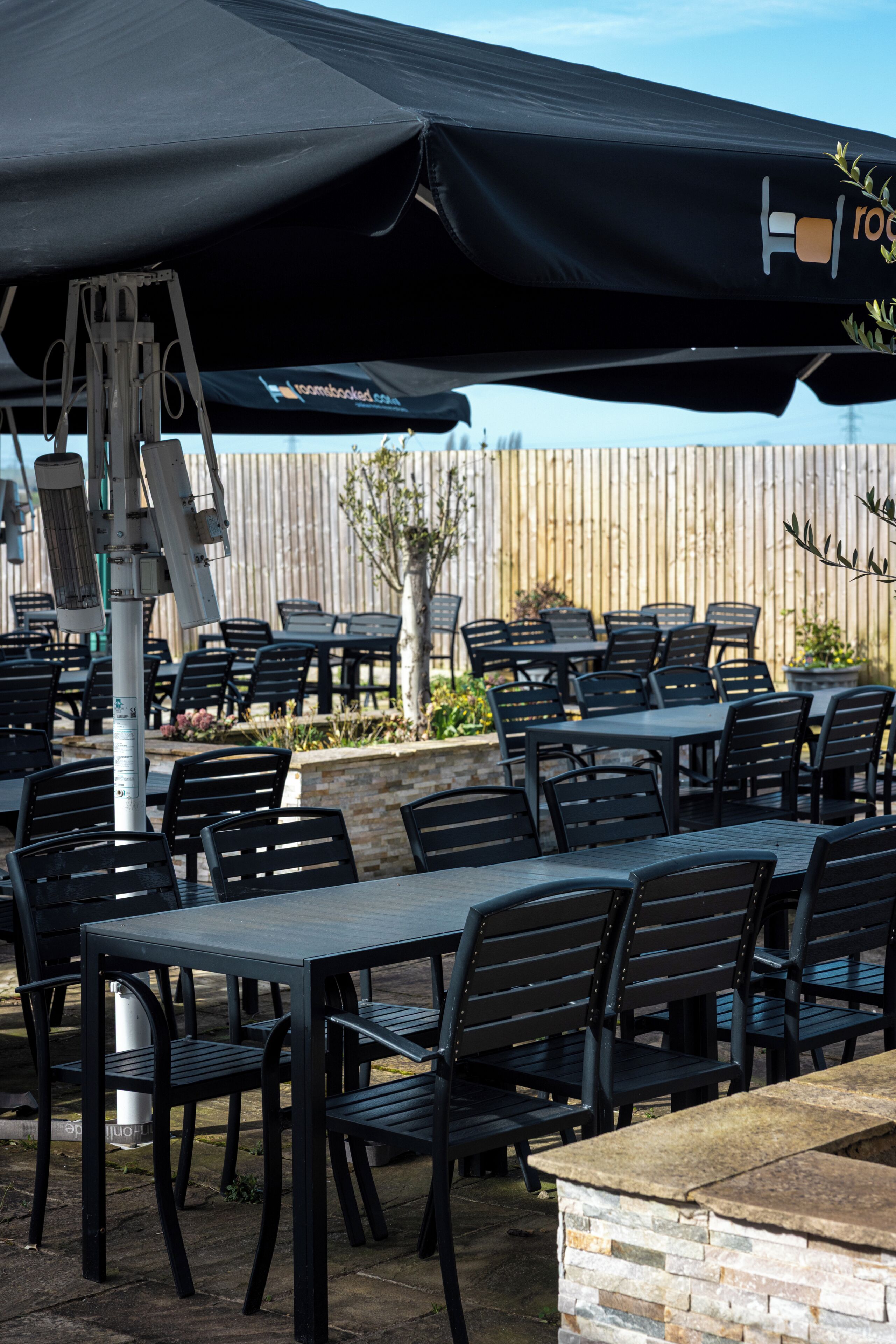 An outdoor patio area with tables and chairs, a wooden fence in the background, and a black umbrella providing shade.