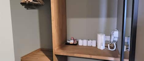 Esquina de una habitación con un estante de madera, un cubo de basura pequeño y un enchufe eléctrico blanco.