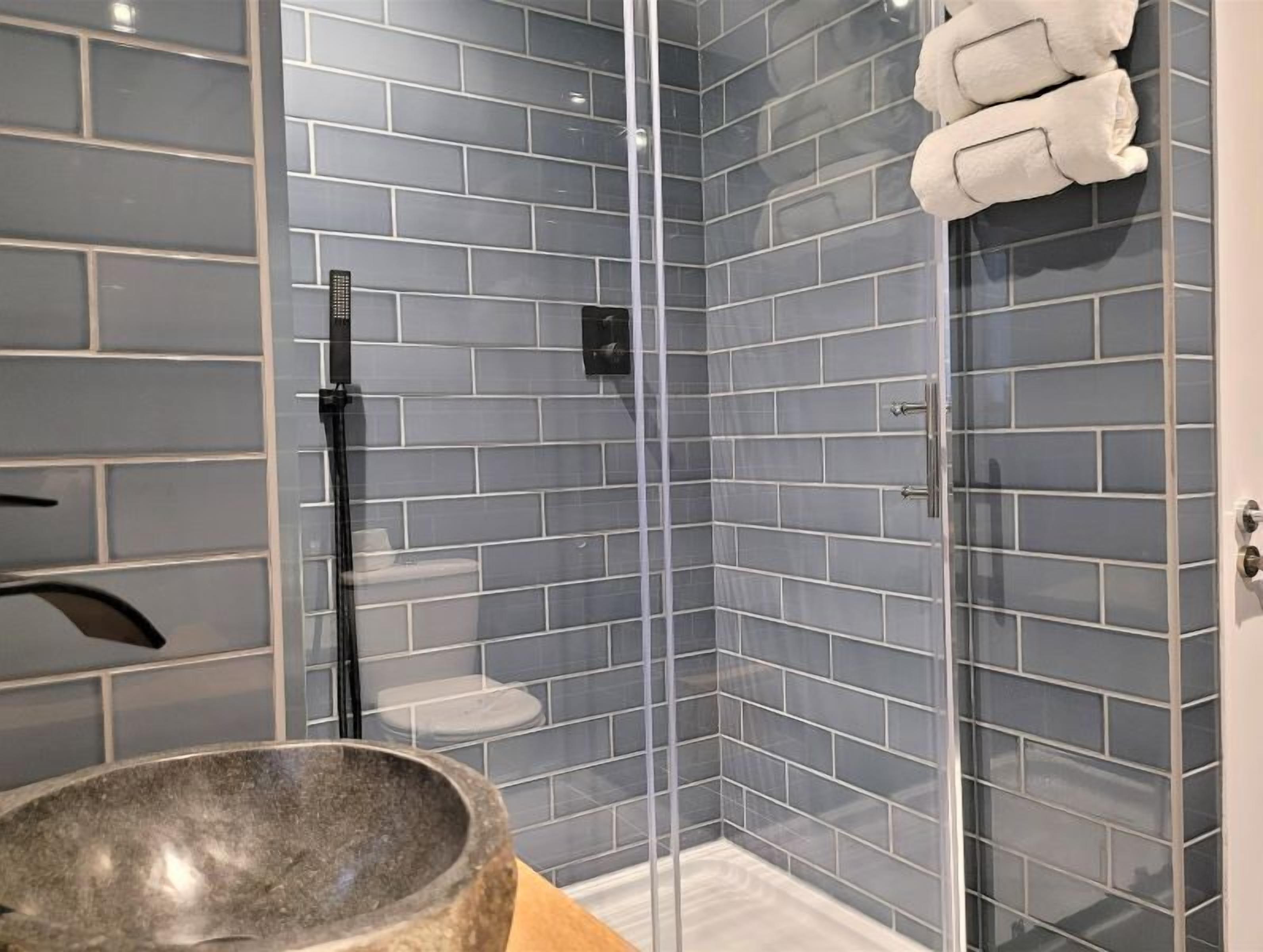 Un baño moderno con paredes de azulejos grises, cabina de ducha de vidrio, lavabo blanco y un perchero para toallas.