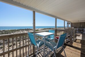 A covered outdoor deck with a view of the beach, wooden furniture, and a white table.