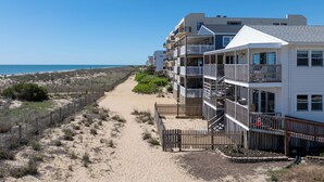 A beachfront property with multiple-story buildings, a sandy path, and a wooden fence. - North OC, charming 2 bedroom oceanfront with Friday to Friday stay (Ocean City)