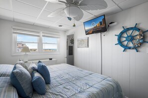 A bedroom with a bed, a television, a ceiling fan, a window, and a wall-mounted ship's wheel.