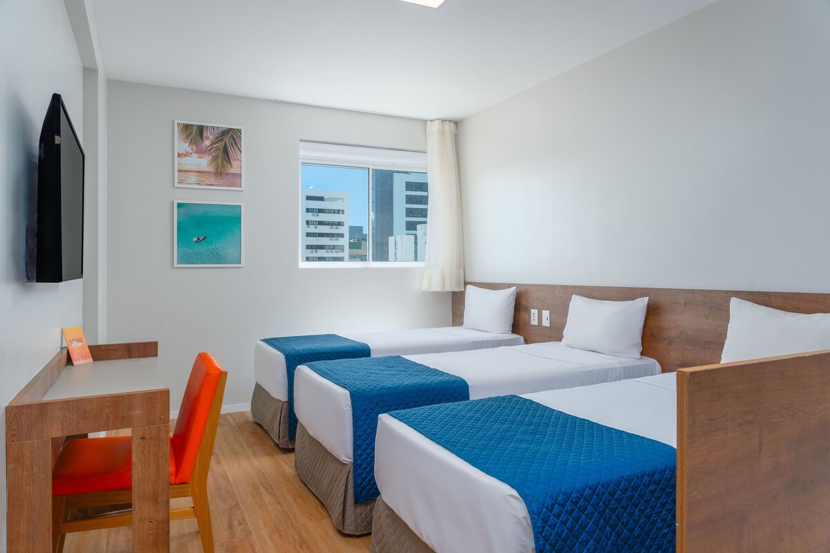 A hotel room with three beds, a desk, a chair, and a TV.