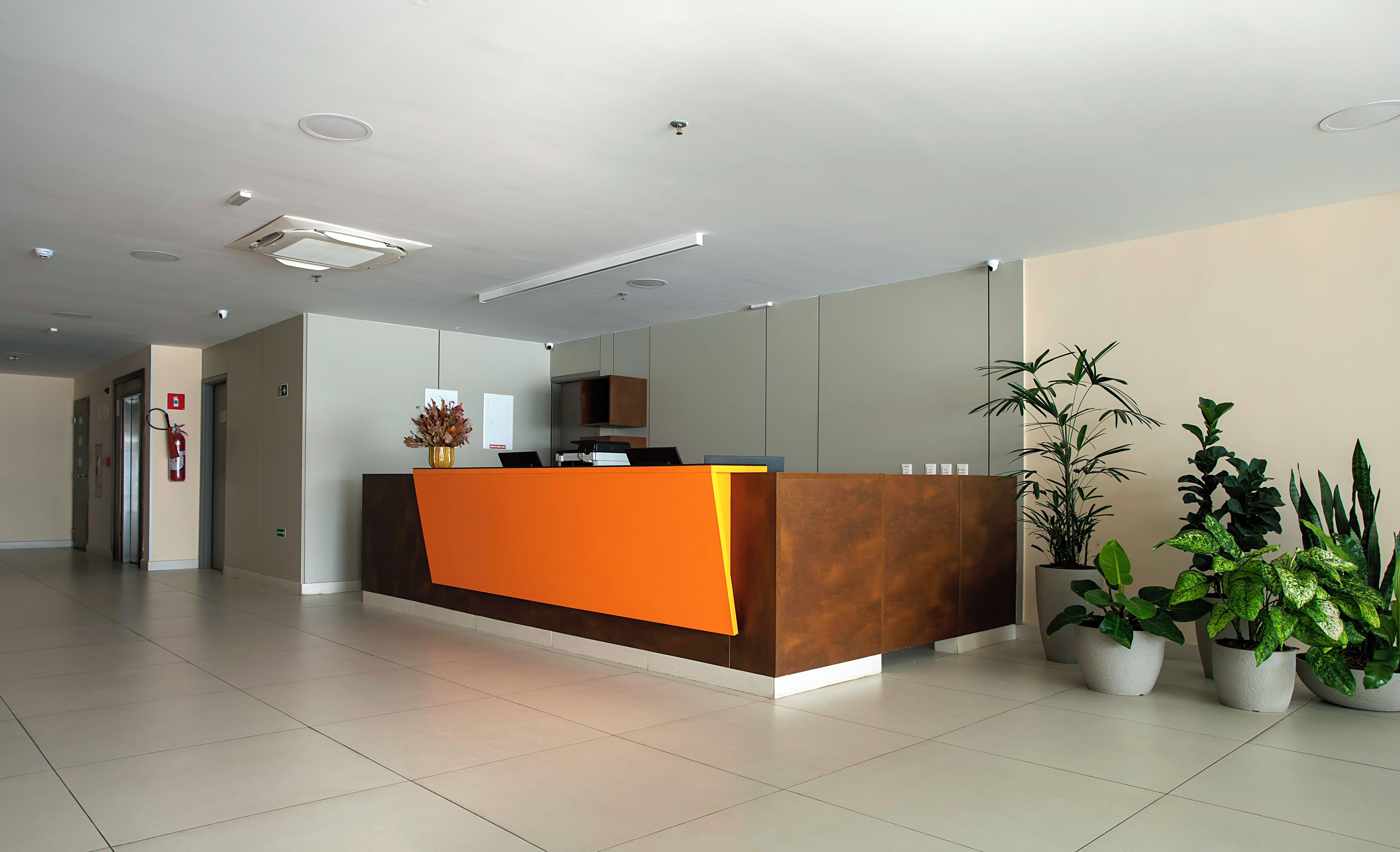 A reception desk with a modern design, a fire extinguisher mounted on the wall, and potted plants around the area.