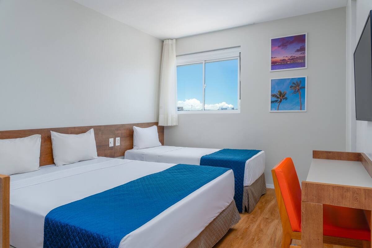 Ein Hotelzimmer mit zwei Betten, einem Schreibtisch und einem Sessel. Ein Fenster bietet Blick auf den Himmel und die Wolken.