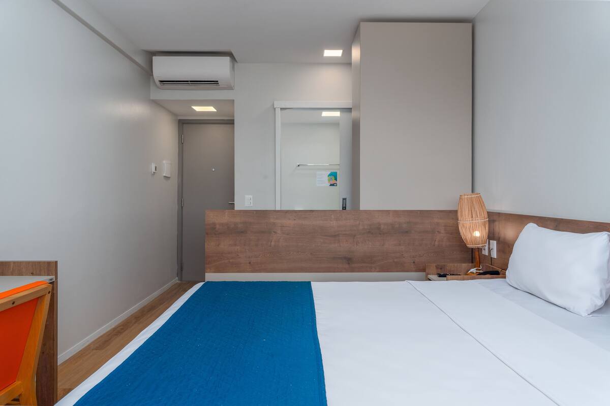 Ein Hotelzimmer mit Holz-Kopfteil, einem Bett mit weißer und blauer Bettwäsche, einer Nachttischlampe und einem kleinen Tisch mit Stuhl.