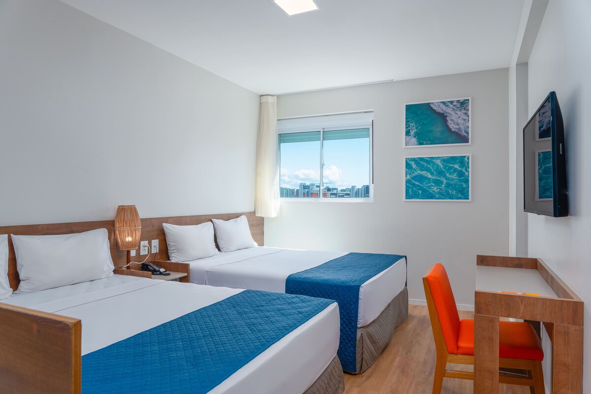A hotel room with two beds, a desk, a chair, and a TV.