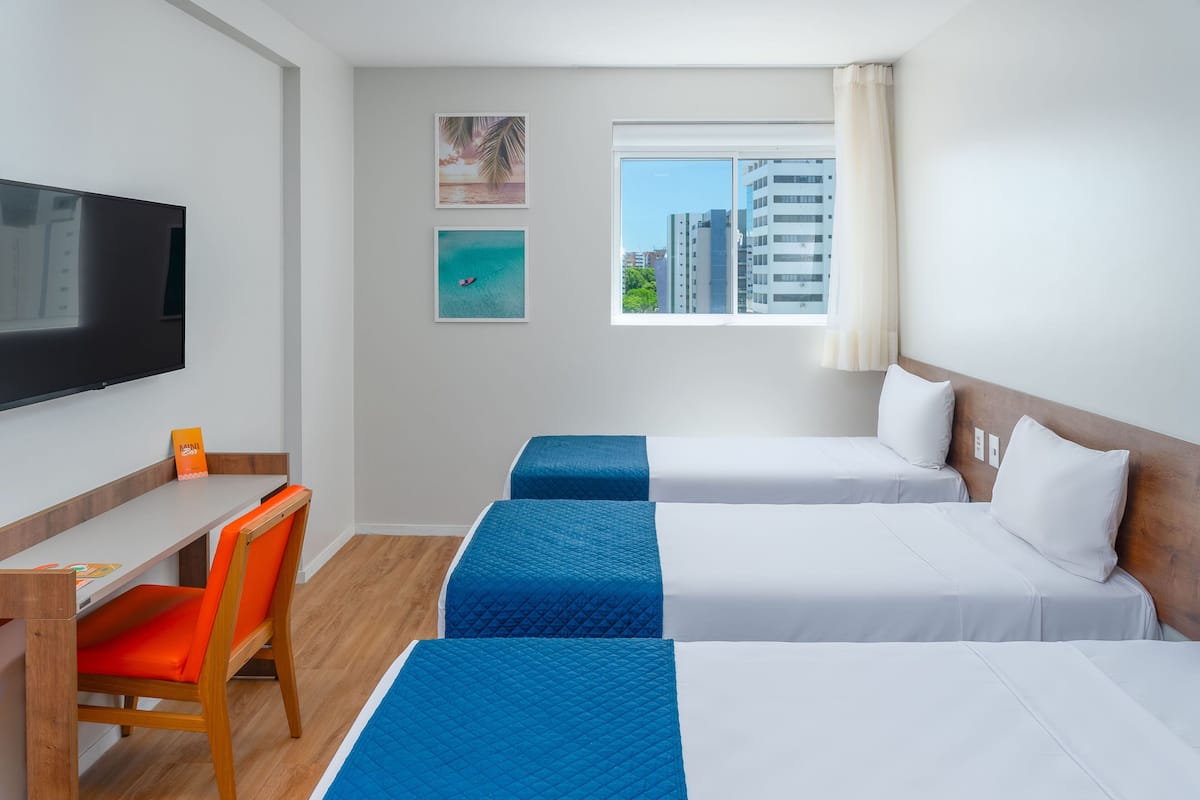 Ein Hotelzimmer mit zwei Betten, einem Schreibtisch, einem Stuhl, einem Fernseher und einem Fenster mit Blick auf die Stadt.