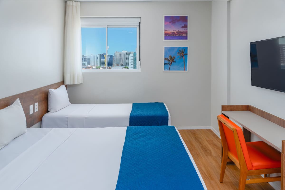 Ein Hotelzimmer mit zwei Betten, einem Schreibtisch, einem Stuhl, einem Fernseher und einem Fenster mit Blick auf die Stadt.