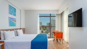 A hotel room with a bed, a desk, a chair, a TV, and large windows offering a city view. - Verano Pajucara By Tropicalis (Maceió)
