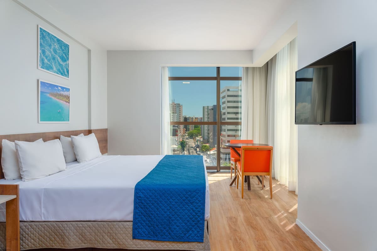 Ein Hotelzimmer mit Bett, Schreibtisch, Stuhl, Fernseher und großen Fenstern mit Blick auf die Stadt.