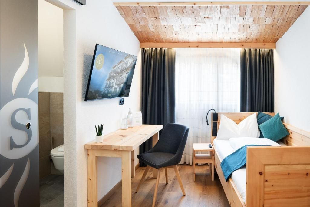 A hotel room with a wooden ceiling, a bed with pillows, a desk with a chair, a TV on the wall, and a window with curtains.