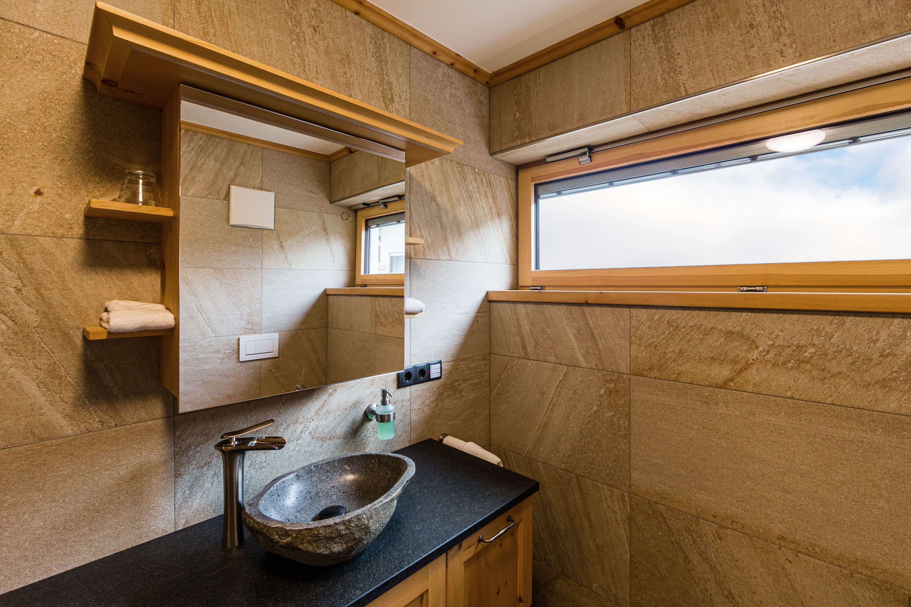A bathroom with a stone sink, wooden cabinets, a mirror, and a window.