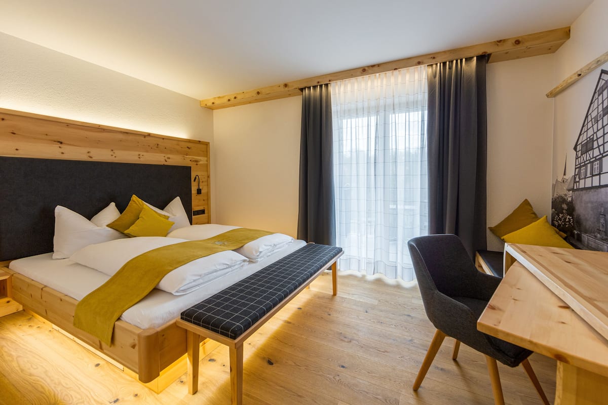 A modern hotel room with a wooden bed, a bench, a dining table, and a large window with curtains.