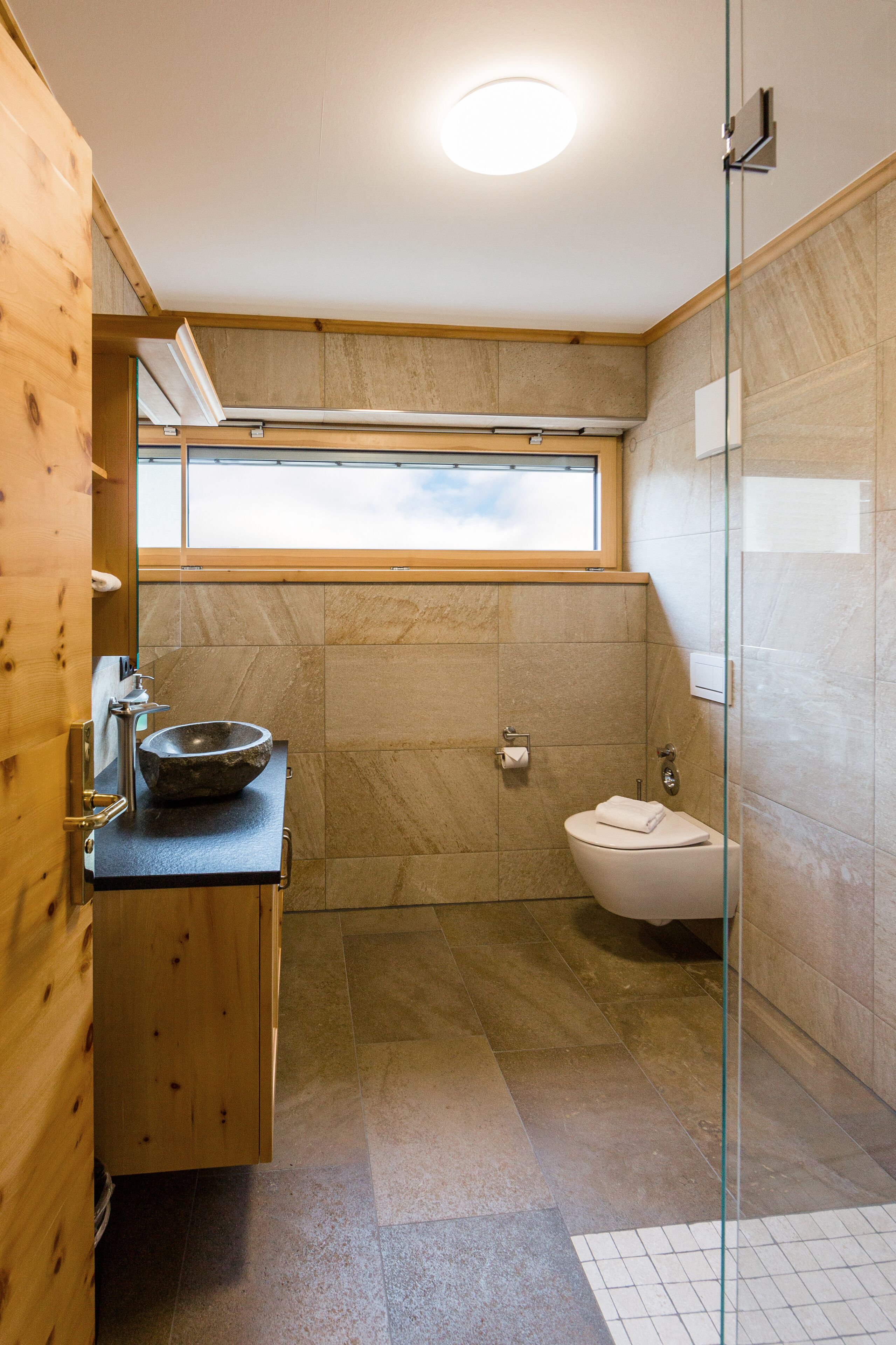 A modern bathroom with a wooden vanity, a glass shower enclosure, and a window above the toilet.
