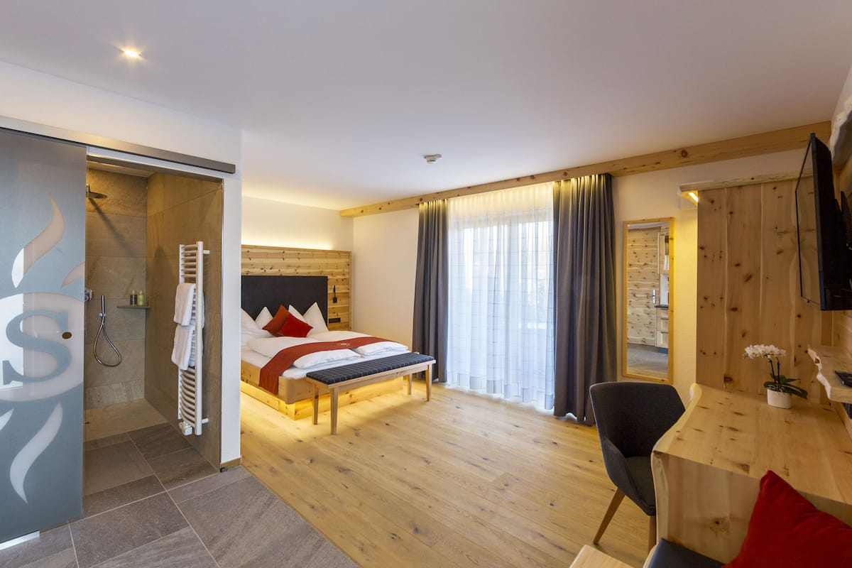 A modern hotel room with a wooden floor, a bed with red and white bedding, a desk with a chair, and a shower area.