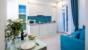 A modern kitchen with white cabinets, a microwave, and a blue tiled backsplash. There is a dining table with a vase of flowers, a bottle of wine, and a basket of bread. A teal sofa is visible in the living area. - Family Apartment in Sorrento Centre (Sorrento)