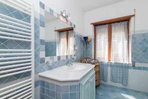 Un baño con un lavamanos blanco, un espejo, una ventana con cortina y suelo de baldosas.
