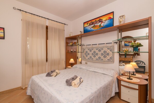 A bedroom with a bed, a nightstand, a mirror, a shelf with decorative items, and a painting on the wall.