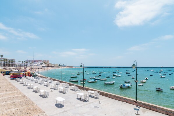 A seaside promenade with rows of white tables and chairs, a beach in the background, and numerous boats on the water.