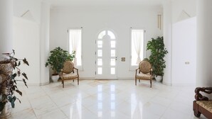 Una habitación amplia y luminosa con piso pulido, dos ventanas grandes con cortinas y dos plantas en macetas.