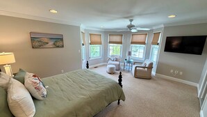 Une chambre à coucher comprenant un lit, un bureau, deux chaises, un ventilateur de plafond et un téléviseur fixé au mur.