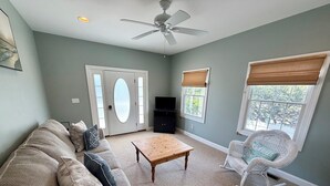 Un salon avec un canapé beige, une table basse en bois, un fauteuil à bascule blanc, un ventilateur de plafond et un téléviseur.