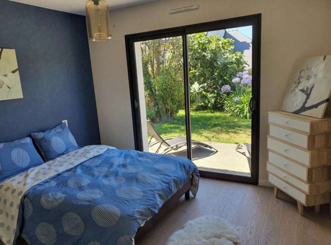 A bedroom with a bed, a dresser, and a sliding glass door leading to an outdoor area with a patio and greenery.