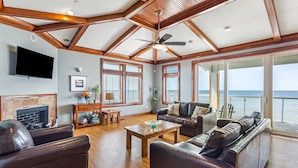 A spacious living room with a fireplace, leather sofas, wooden flooring, and large windows offering a view of the ocean. - WV31 Sky Surfer by First Flight Rentals (Rodanthe)