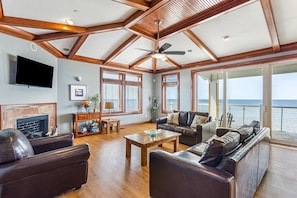 A spacious living room with a fireplace, leather sofas, wooden flooring, and large windows offering a view of the ocean. - WV31 Sky Surfer by First Flight Rentals (Rodanthe)
