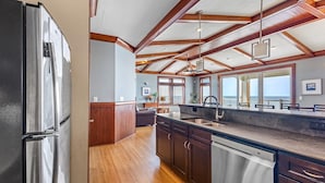 A kitchen with wooden cabinets, a stainless steel refrigerator, and a large window overlooking the ocean. - WV31 Sky Surfer by First Flight Rentals (Rodanthe)