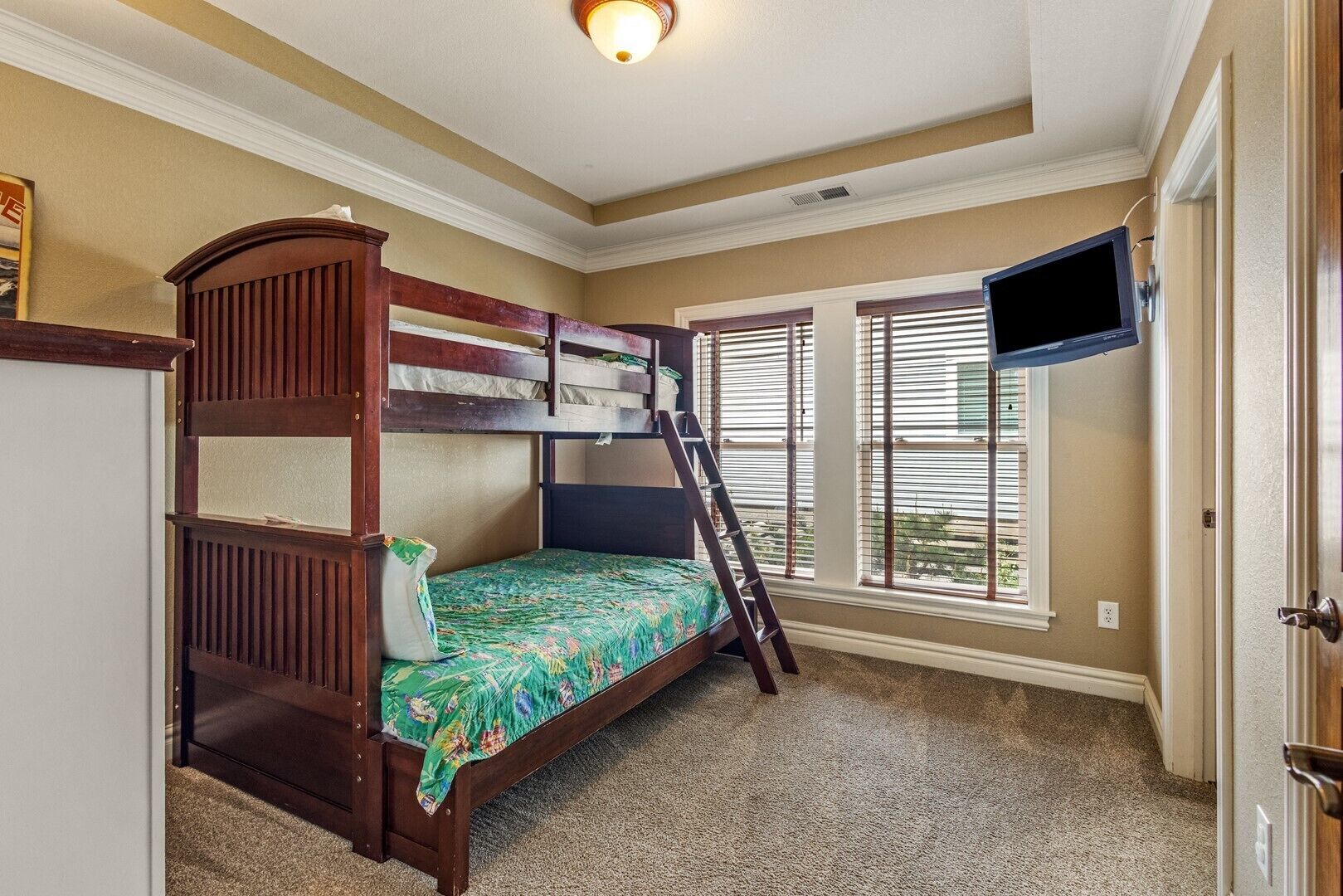A bunk bed room with a TV mounted on the wall, carpeted floor, and a window with blinds.