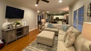 Un salon comprenant un canapé d’angle, un téléviseur fixé au mur, un ventilateur de plafond et un meuble de rangement en bois pour les appareils multimédias.