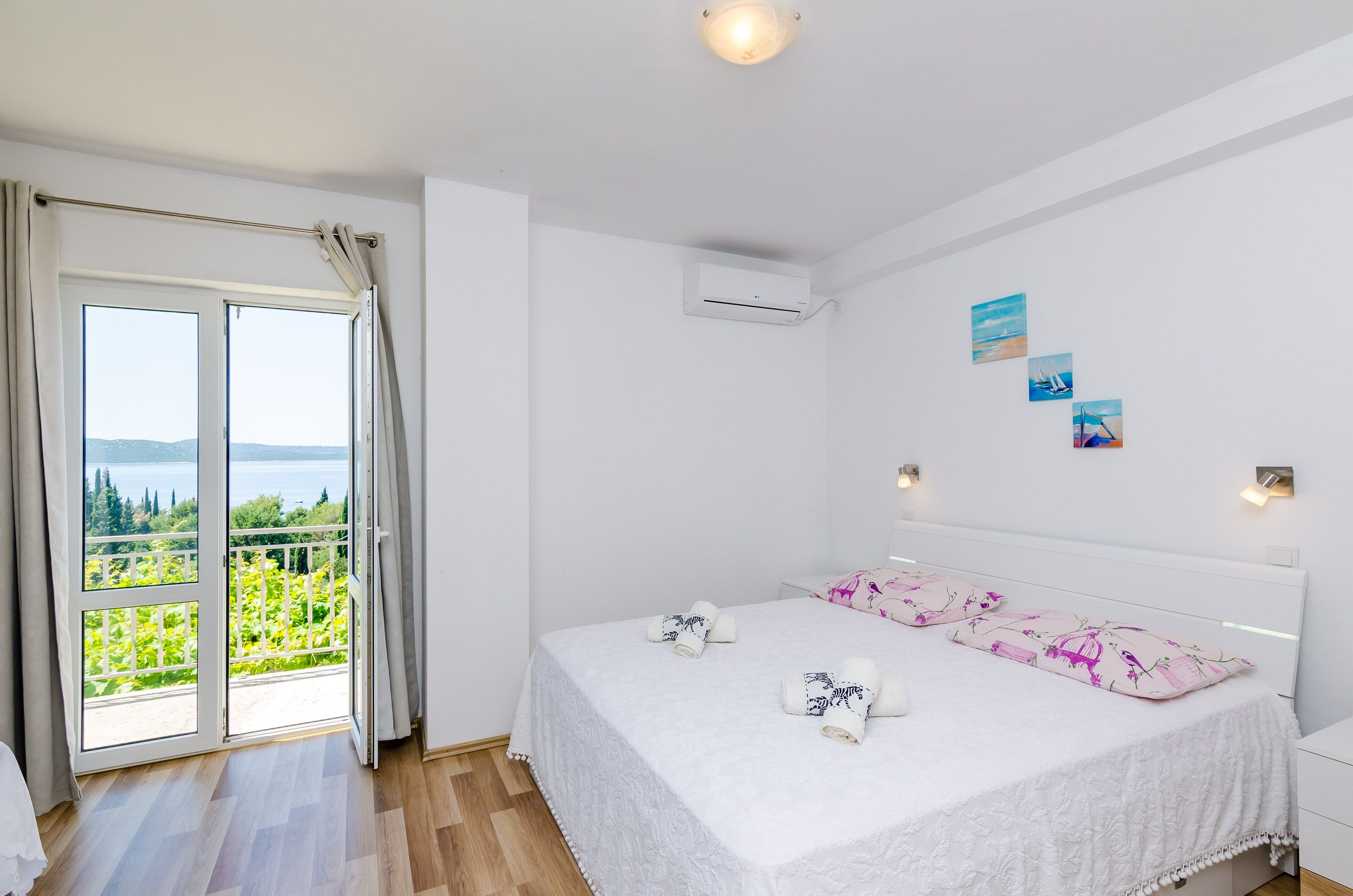 A bedroom with a large window, a bed with white bedding, and a view of greenery and a body of water.