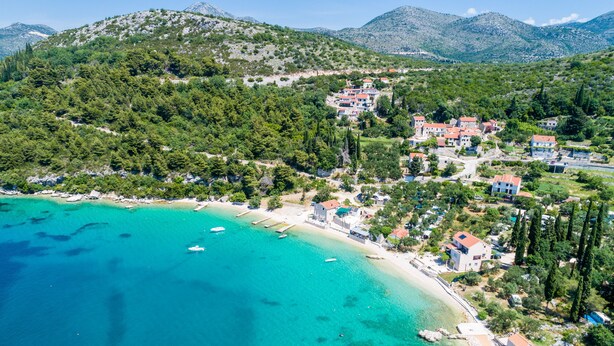 Luftaufnahme eines Küstdorfes mit Gebäuden, einem Strand und klarem türkisblauem Wasser.