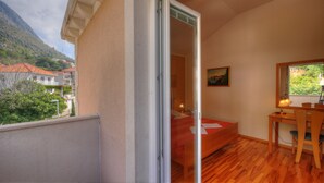 A bedroom with a wooden floor, a bed, a desk, and a chair. There is a window with a view of buildings and trees. - Apartment Villa Molina (Zupa dubrovacka)