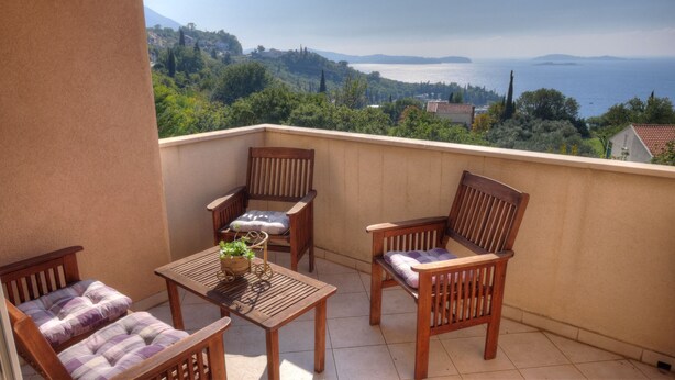 A balcony with wooden furniture, including two chairs and a table, overlooking a scenic view of a body of water and hills. - Apartment Villa Molina (Zupa dubrovacka)